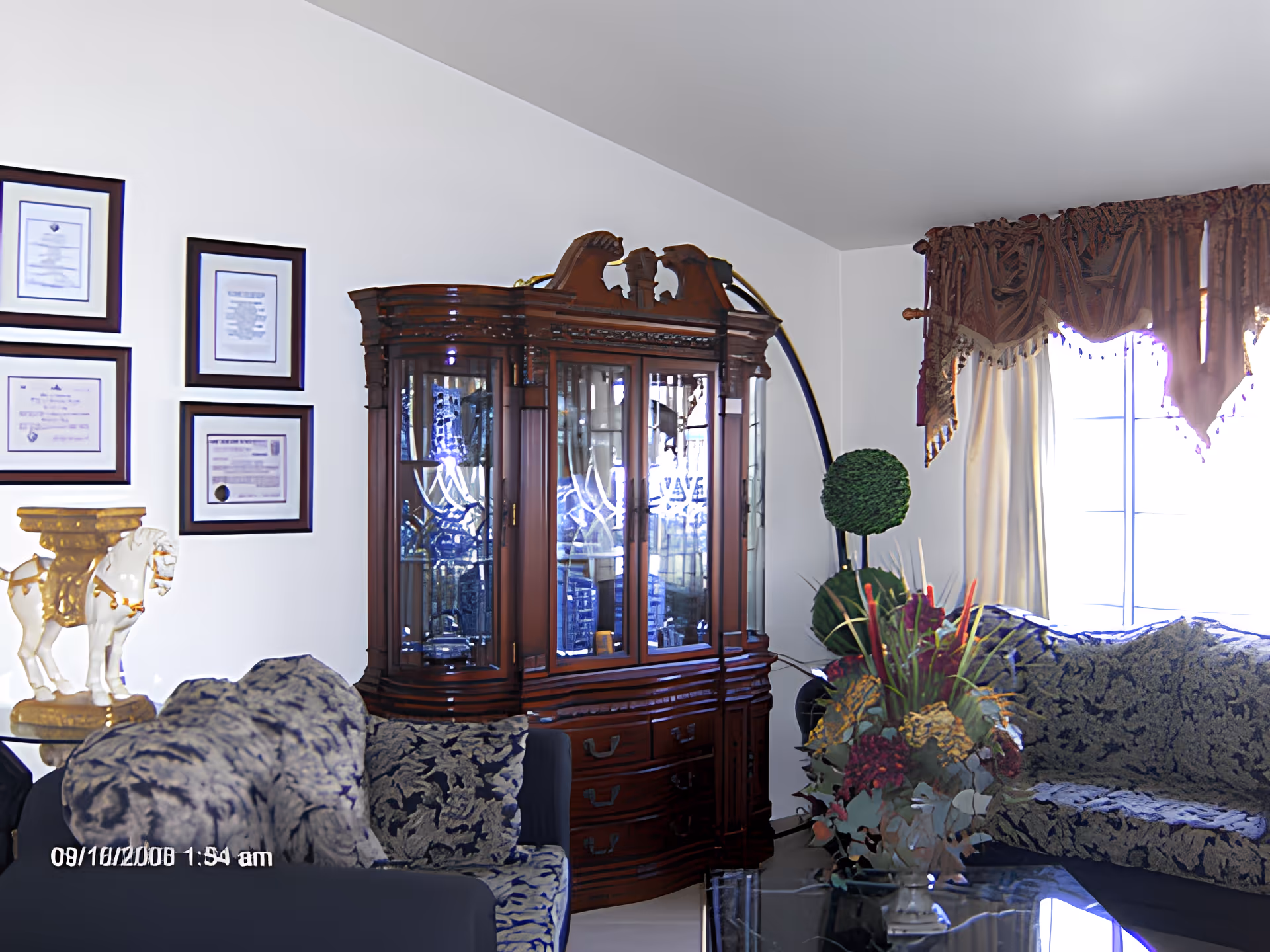 Sunlit living room with patterned sofas, an ornate wooden china cabinet, a floral centerpiece, and framed certificates on the wall.