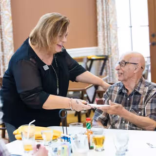 A woman serving a dessert to an elderly man seated at a dining table in a communal dining area. The man is smiling and wearing glasses and a plaid shirt. The woman is smiling and wearing a black top. The table has various glasses, condiments, and utensils on it, and the background shows windows with curtains.