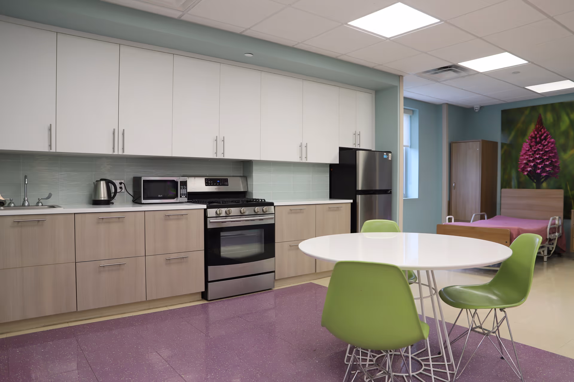 A modern kitchenette and dining area with white and wood cabinets, stainless steel appliances, a round table with green chairs, and a bed with floral artwork in the background.
