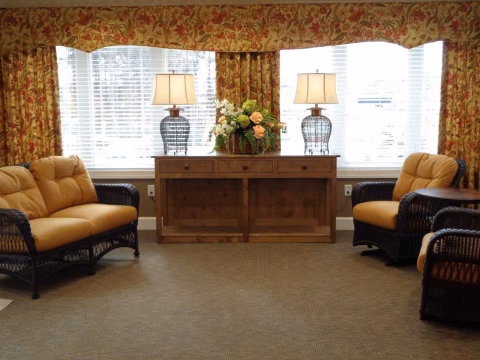 A cozy sitting area with two cushioned wicker armchairs and a matching loveseat arranged around a wooden console table. The table holds two decorative lamps and a floral arrangement. Behind the table are large windows with white blinds and floral-patterned curtains.