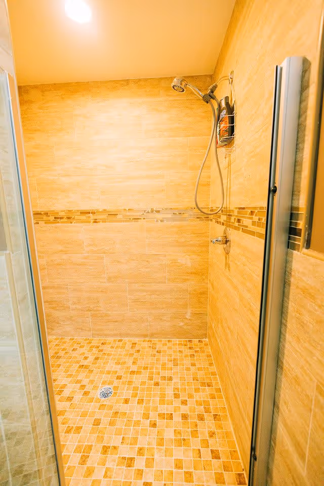 A tiled shower area with beige and brown mosaic floor tiles and beige wall tiles featuring a decorative horizontal strip. The shower has a handheld showerhead attached to the wall and a small metal rack holding shower products. The shower is enclosed with a glass door.