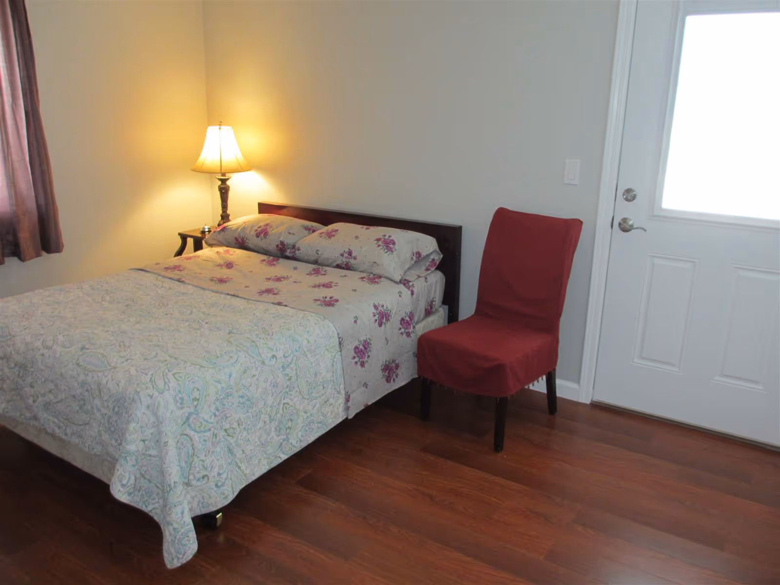 Small bedroom with a floral-patterned bed, a lit bedside lamp, a red-covered chair, and a door to the right.
