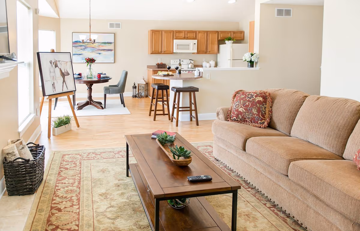 A bright and cozy living room area with a beige sofa adorned with patterned cushions, a wooden coffee table with small potted plants and a remote control, and a decorative rug. In the background, there is a small dining area with a round table set for two, an easel with artwork, and a kitchen with wooden cabinets, a microwave, refrigerator, and two bar stools at a counter.
