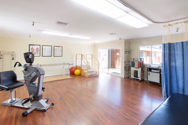 A spacious rehabilitation or physical therapy room with wooden flooring, a stationary exercise bike, parallel bars for walking practice, therapy balls, a small set of stairs for mobility exercises, anatomical charts on the wall, and a blue privacy curtain partially visible on the right side.
