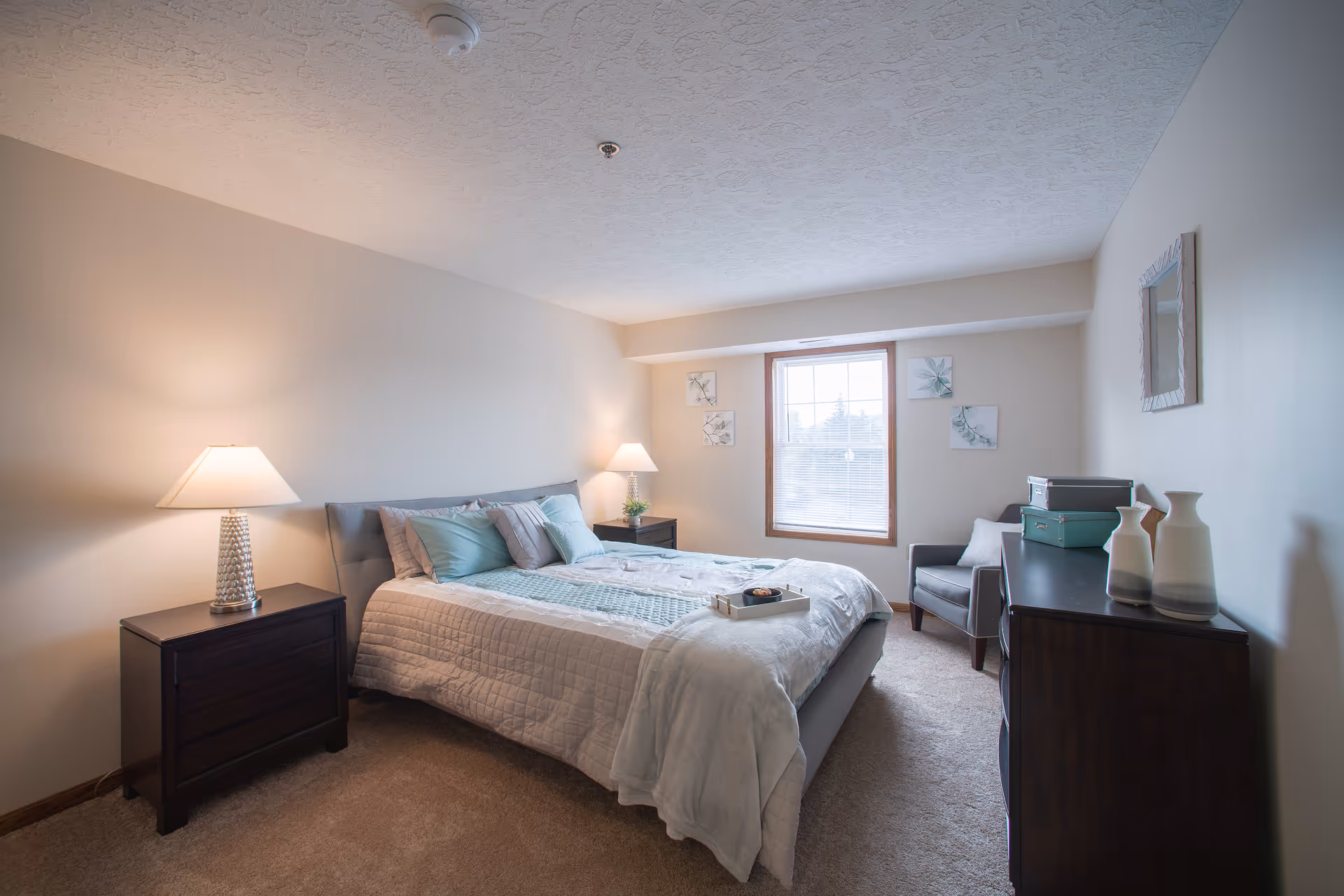 A cozy bedroom with a large bed covered in light-colored bedding and several pillows. There are two bedside tables with lamps on either side of the bed. A window with blinds lets in natural light, and three framed botanical prints hang on the wall around the window. A gray armchair is positioned near the window, and a dark wooden dresser with decorative vases and storage boxes sits against the opposite wall. The room has beige walls and carpeted flooring.