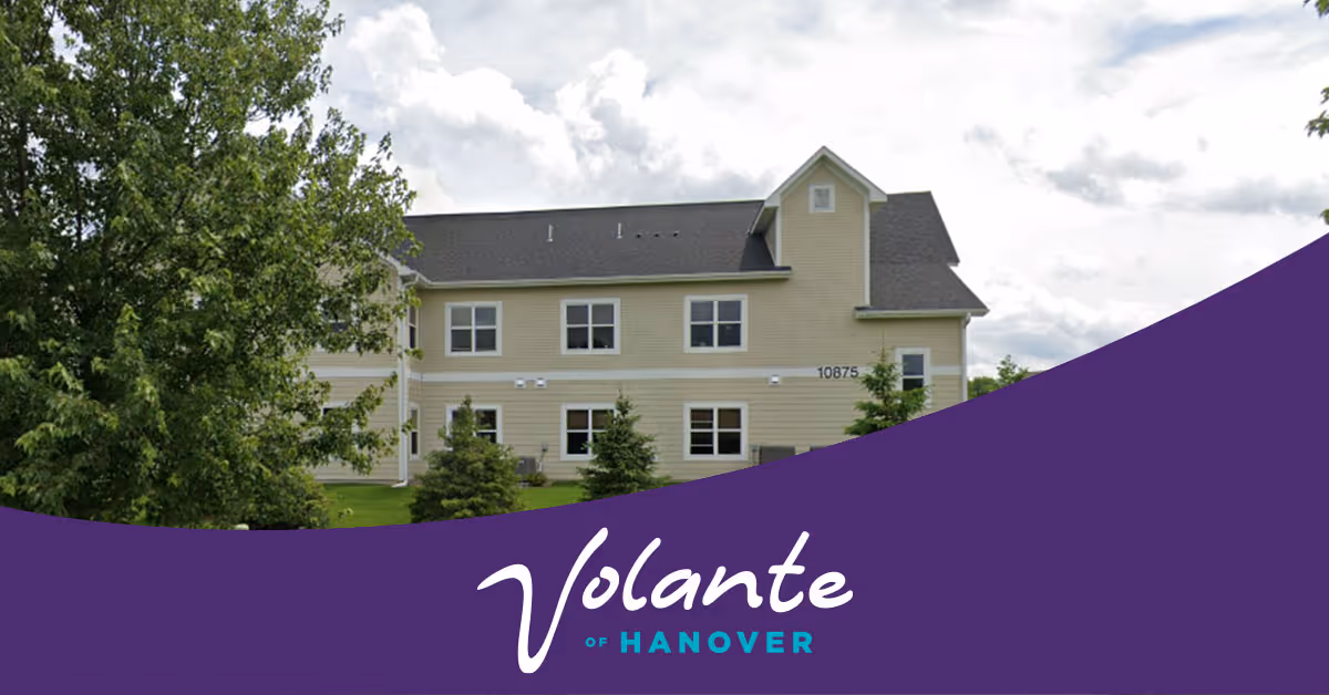 Exterior view of a two-story beige senior living facility building with multiple windows, surrounded by green trees and grass under a partly cloudy sky. The bottom portion of the image has a purple overlay with the text 'Volante of Hanover'.