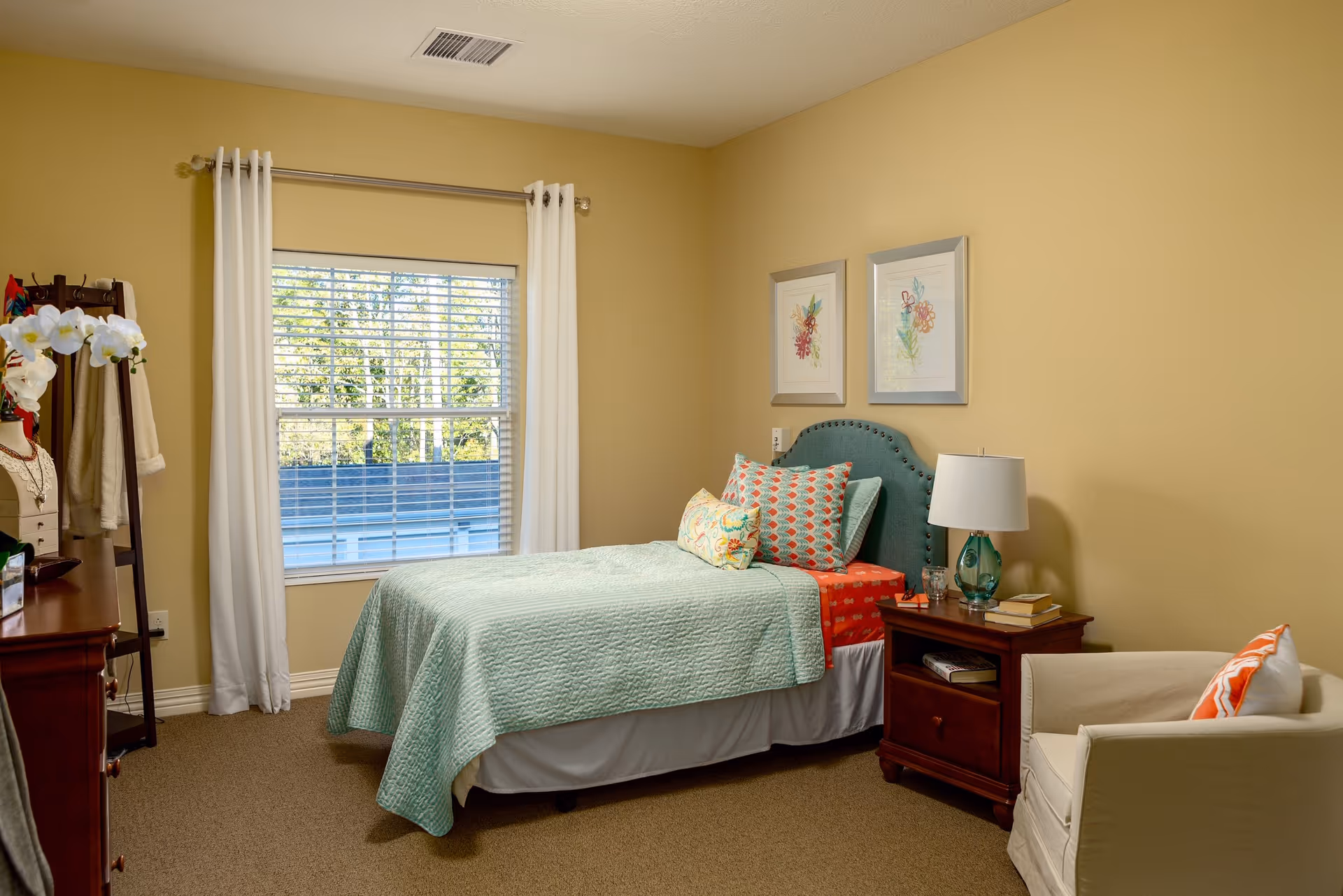 A cozy bedroom with a single bed covered in a light blue quilt and colorful pillows. There is a wooden nightstand with a lamp, books, and a glass of water next to the bed. A beige armchair with an orange and white pillow is placed beside the nightstand. The room has beige walls, a window with white curtains, and two framed floral artworks hanging above the bed. A wooden dresser and a coat rack with clothes are visible on the left side.