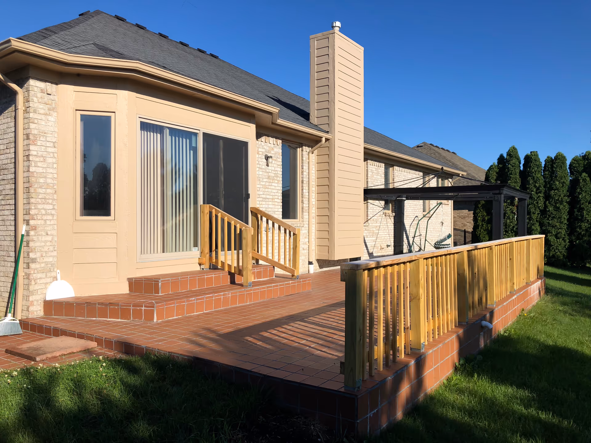 Outdoor view of a single-story building with beige brick walls and a dark shingled roof. There is a tiled patio with wooden railings and steps leading to a sliding glass door. The patio is adjacent to a grassy lawn and there are tall evergreen trees in the background under a clear blue sky.