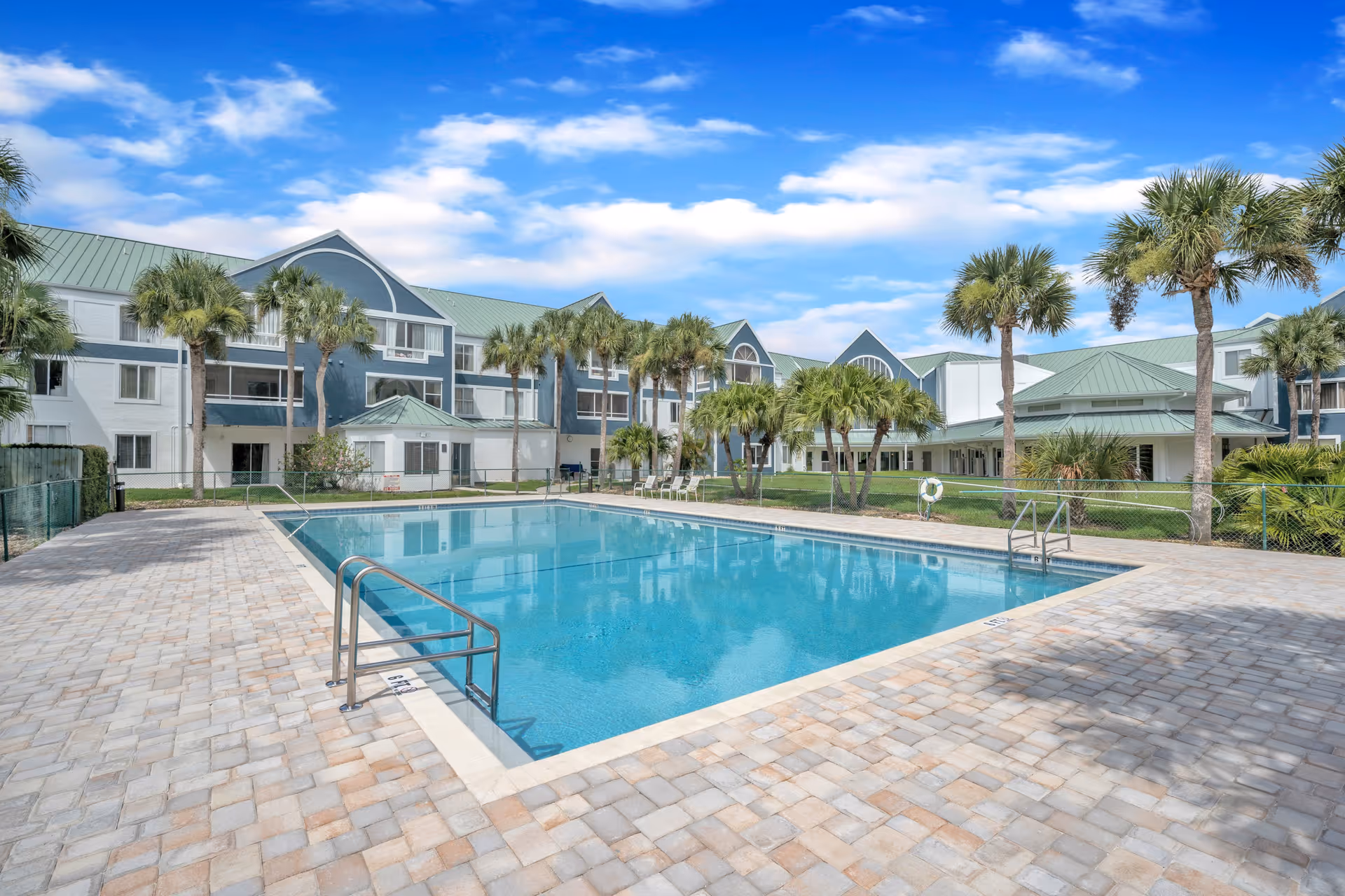 Outdoor swimming pool with a paver deck, palm trees, and a multi-story senior living building under a blue sky.