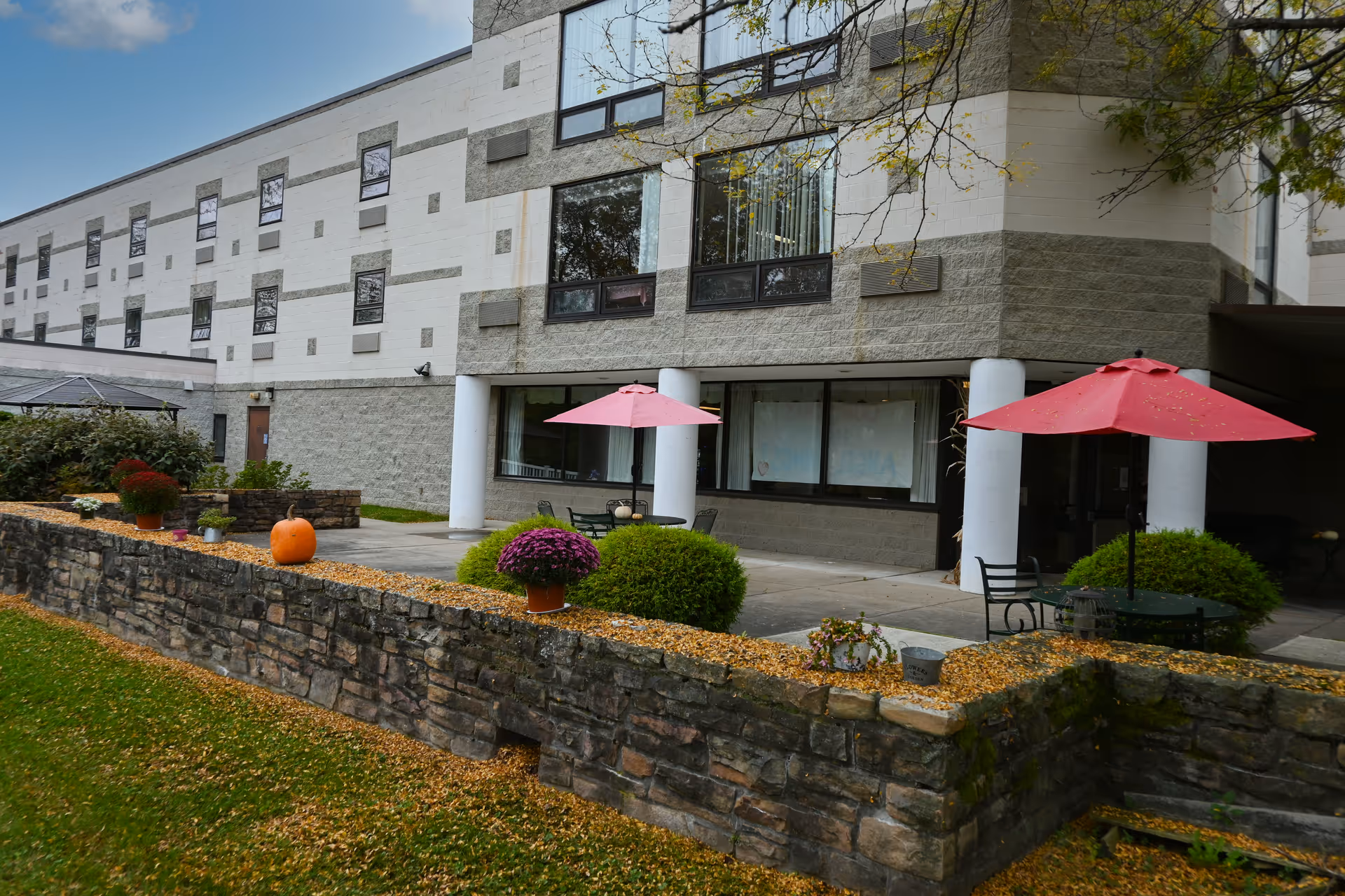 Outdoor patio area of a senior living facility with stone walls, potted plants, a pumpkin, and tables with red umbrellas. The building has large windows and white pillars supporting an overhang.