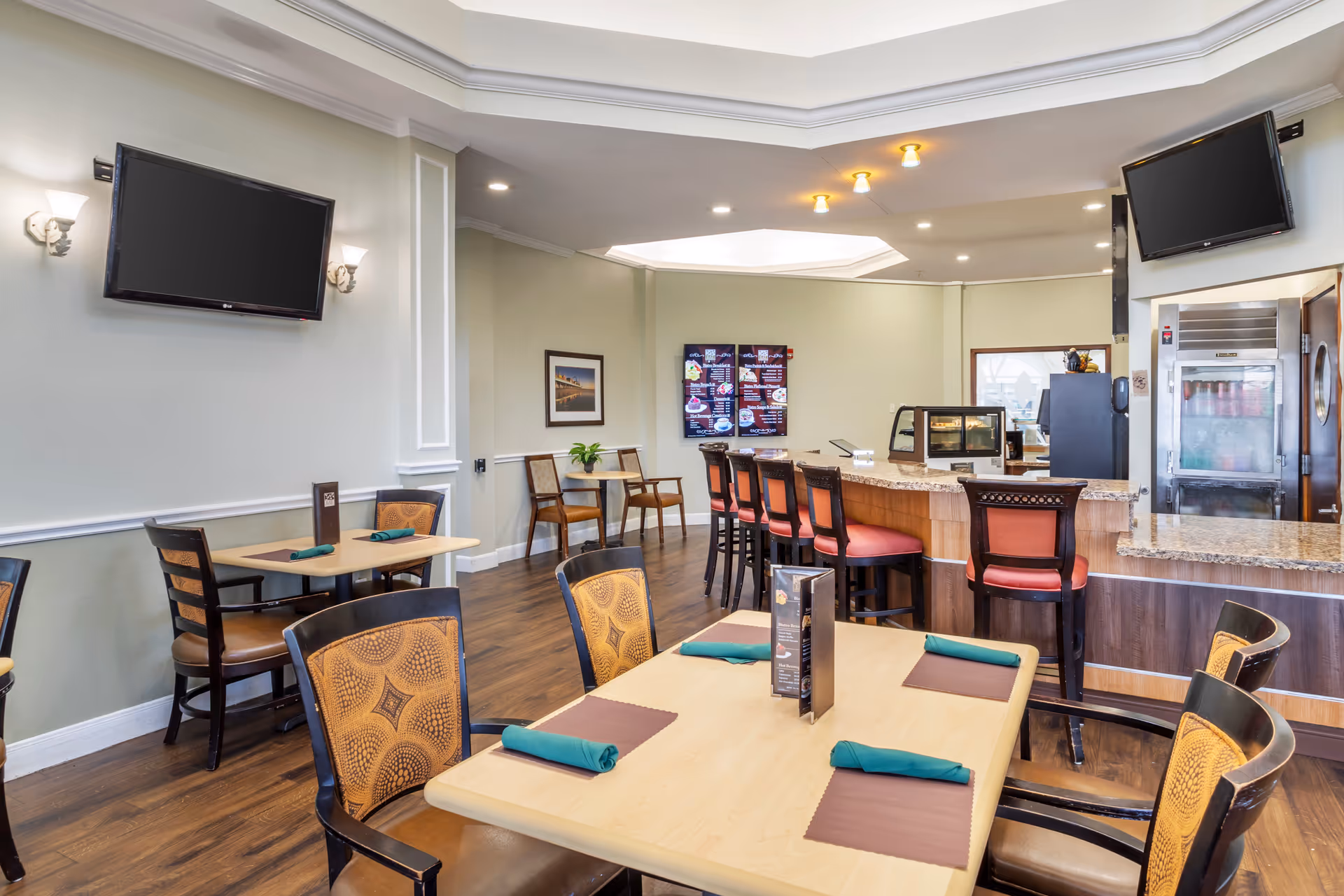 Interior view of a dining area in a senior living facility with wooden tables and chairs, some tables set with green napkins and brown placemats. There is a counter with high chairs, two wall-mounted flat-screen TVs, a digital menu board, and a refrigerator in the background.