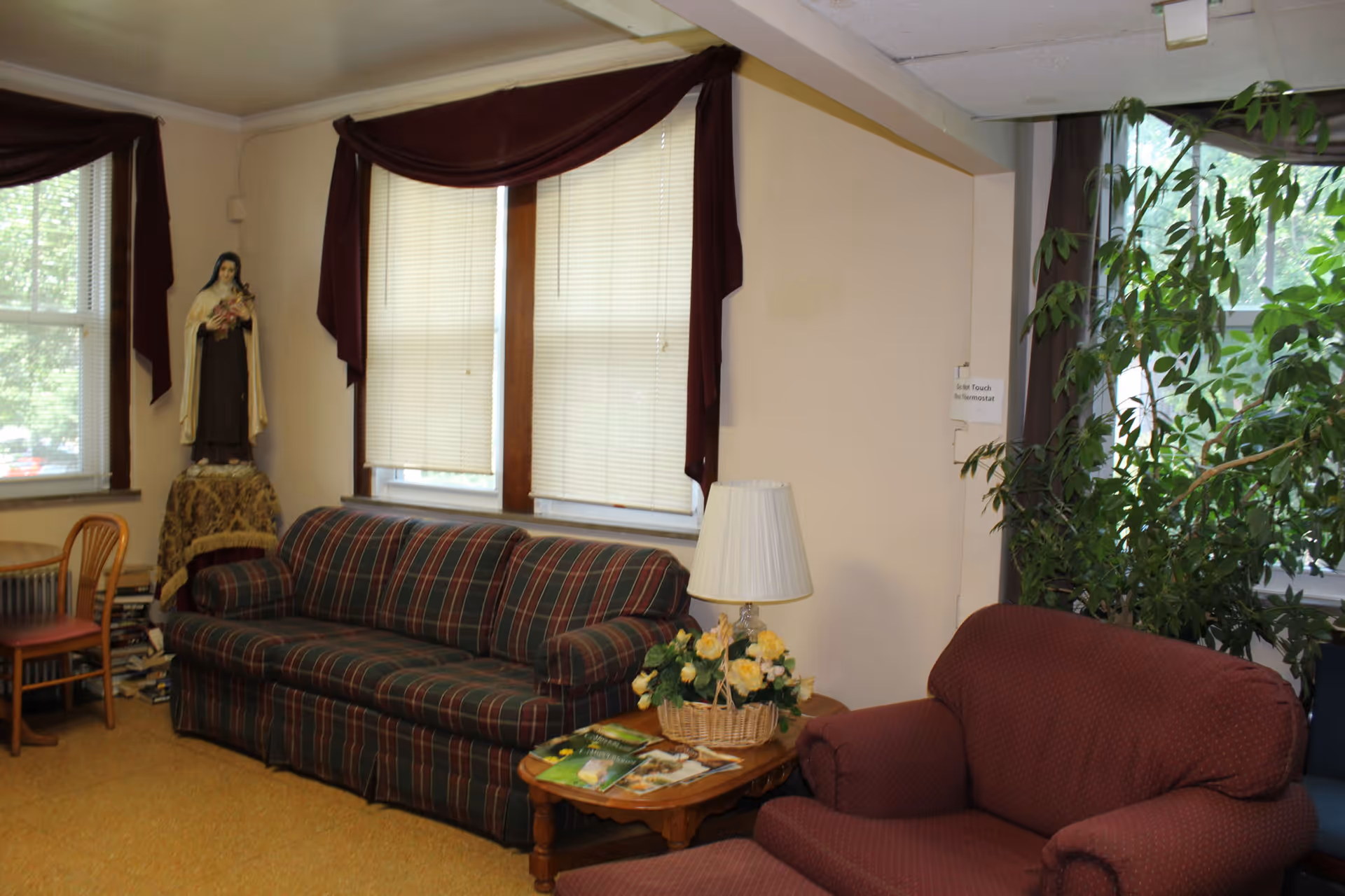 A cozy living room area with a plaid sofa and a maroon armchair. There is a wooden side table with a lamp and a basket of yellow flowers. A statue of a religious figure stands on a small table covered with a cloth near the window. The windows have blinds and maroon valances. A large green plant is placed near another window.