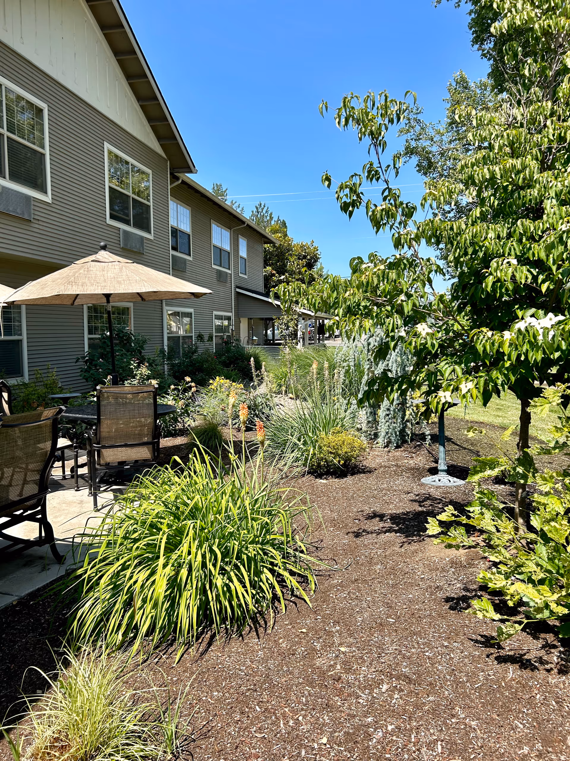 Outdoor courtyard and landscaped garden beside a two-story senior living building with patio seating and an umbrella.
