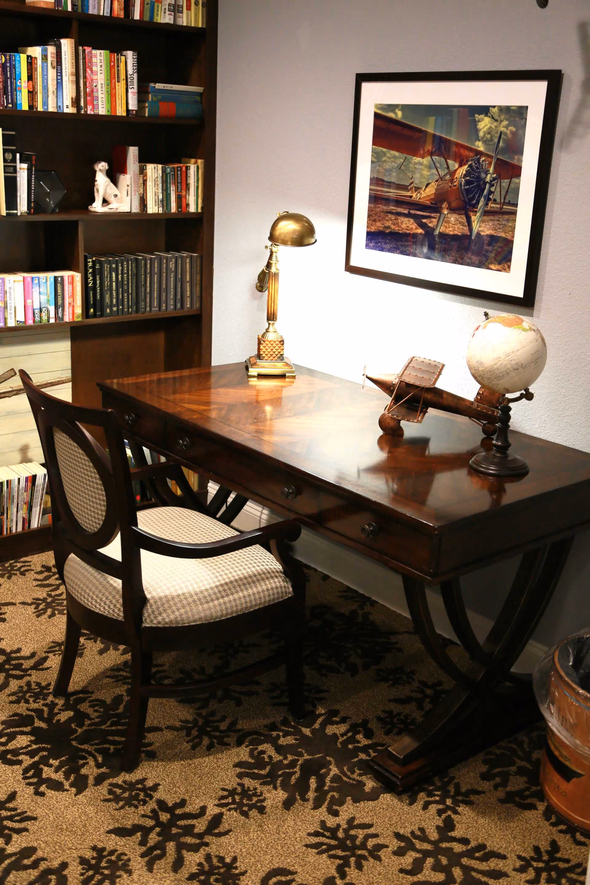 A cozy study area with a wooden desk and a cushioned chair on a patterned carpet. On the desk, there is a vintage-style brass lamp, a small model airplane, and a globe. Behind the desk is a bookshelf filled with books and a small white dog figurine. A framed picture of an old airplane hangs on the wall above the desk.
