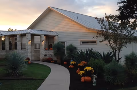 Exterior view of a single-story building with white siding and a metal roof at sunset. A curved concrete pathway leads to the entrance, which is decorated with pumpkins and yellow flowers. Various green shrubs and plants line the pathway and the building.