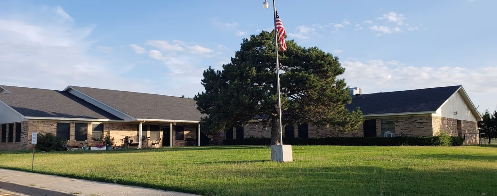 Exterior view of a single-story brick building with a covered porch, surrounded by a well-maintained lawn and a large tree near a flagpole with an American flag, under a partly cloudy sky.