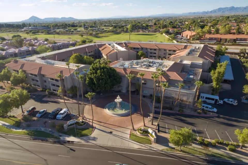 Aerial view of the Grand Court of Mesa senior living facility showing a large building with a red-tiled roof, a circular driveway with a central fountain, several palm trees, parked cars, and surrounding greenery with mountains in the background.