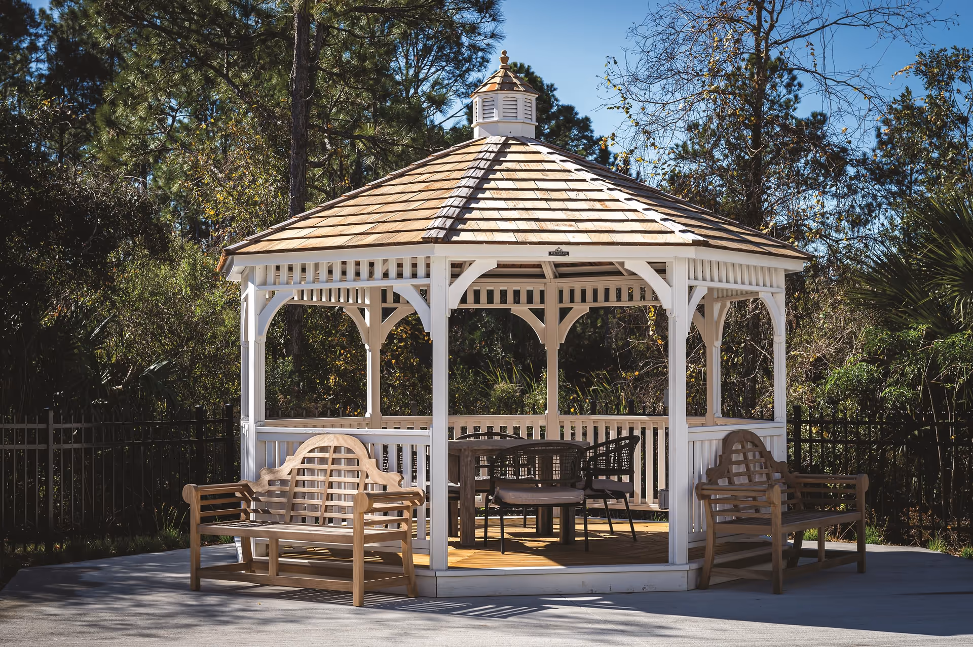 White wooden gazebo with benches and chairs set outdoors among trees.