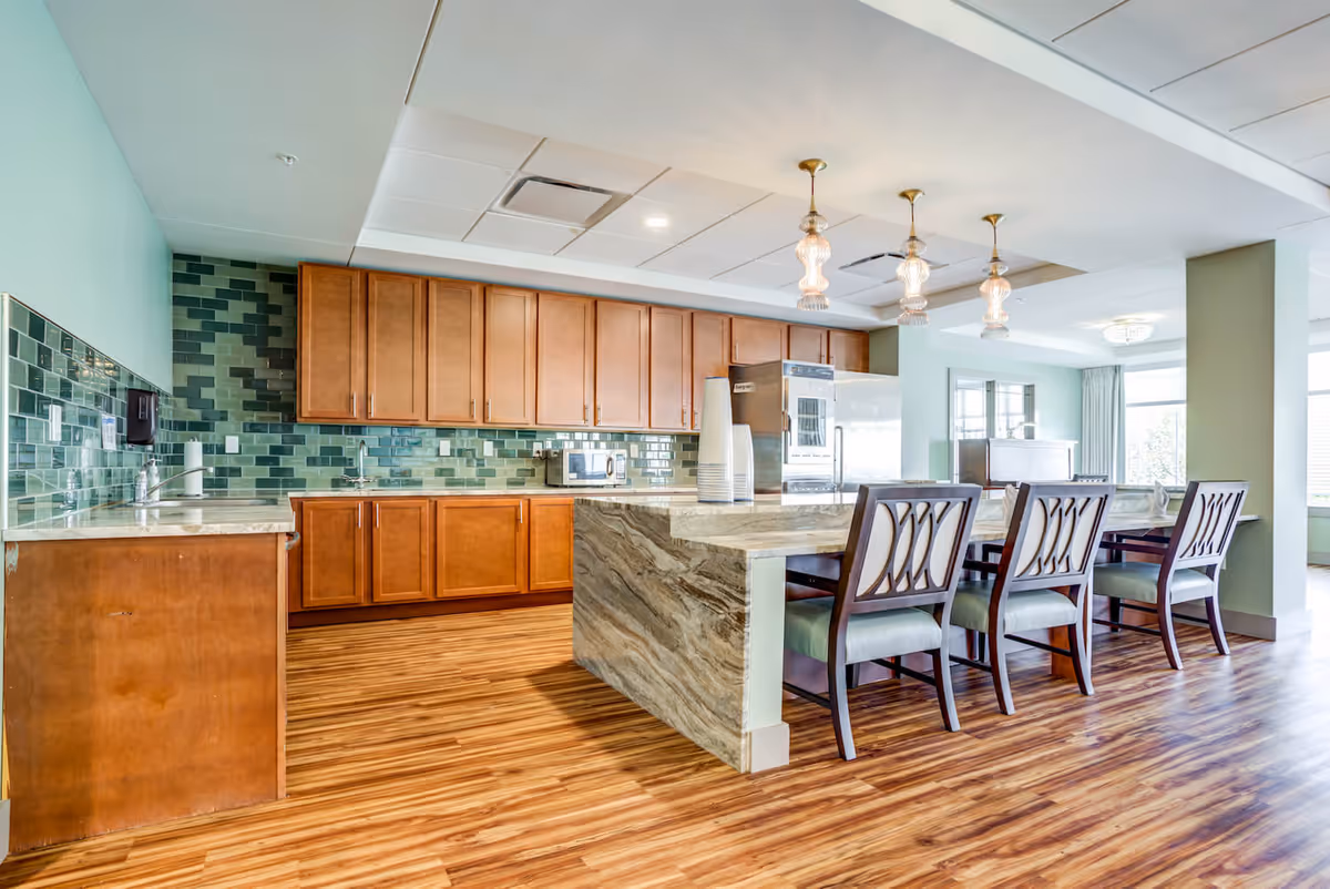 A bright and spacious kitchen area with wooden cabinets, green tiled backsplash, and a marble countertop island with four wooden chairs. The floor is wood-patterned, and there are three pendant lights hanging from the ceiling. Large windows allow natural light to fill the space.