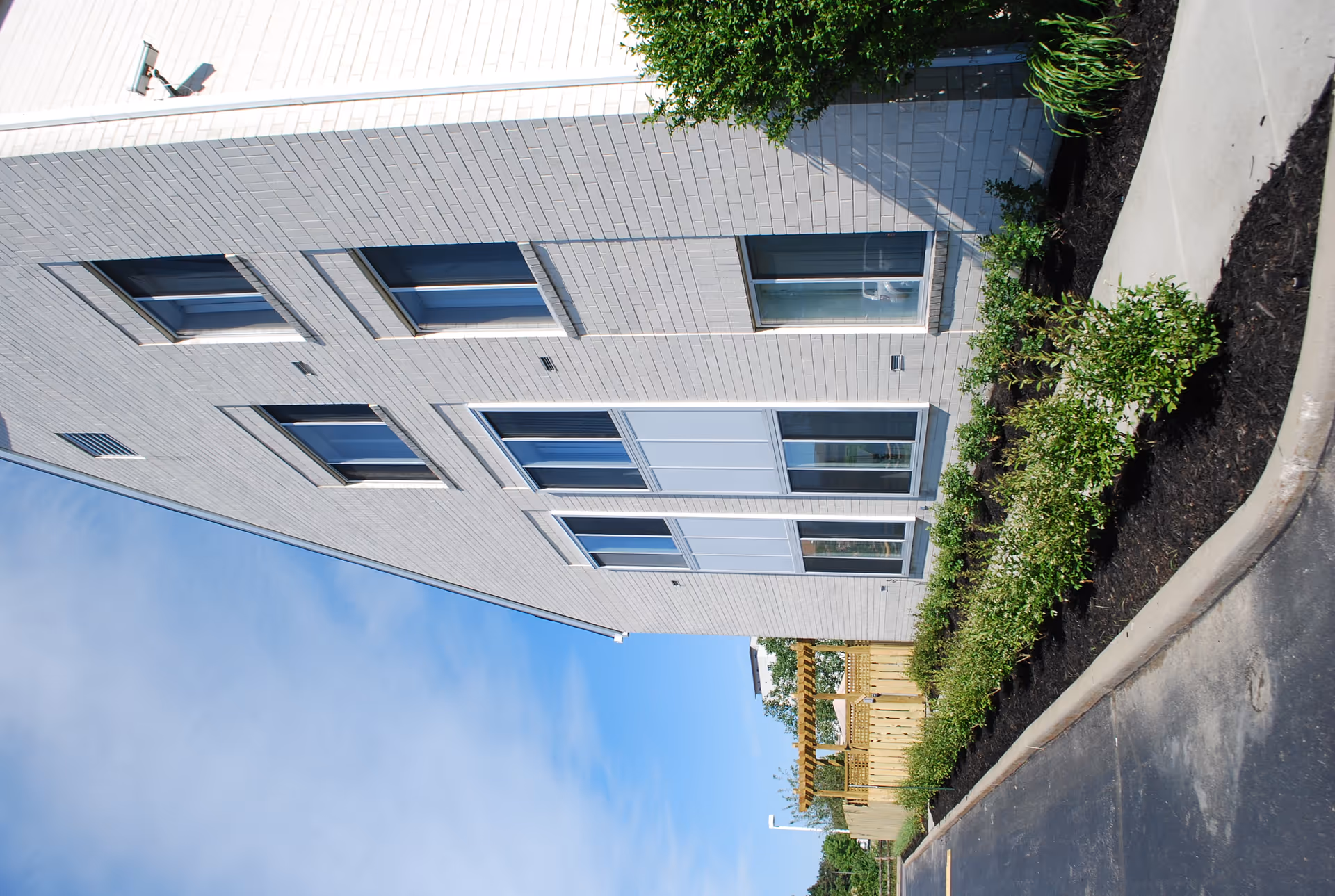 Exterior view of a light-colored brick building with multiple windows, landscaped shrubs, a sidewalk and a blue sky.
