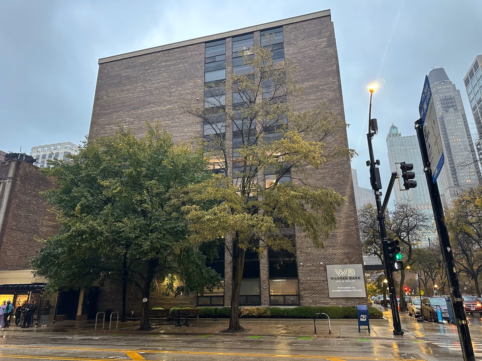 Exterior view of a multi-story brick building with trees in front on a rainy day. The building has a sign that reads 'Warren Barr Gold Coast'. Cars and streetlights are visible on the street beside the building.