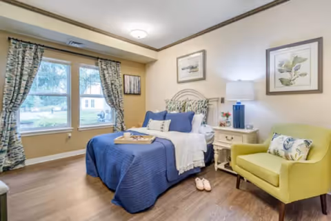 A cozy bedroom with a bed dressed in blue bedding and white pillows, a tray on the bed, a yellow armchair with a floral pillow, a nightstand with a lamp and small decorations, two framed pictures on the wall, and a window with patterned curtains letting in natural light.