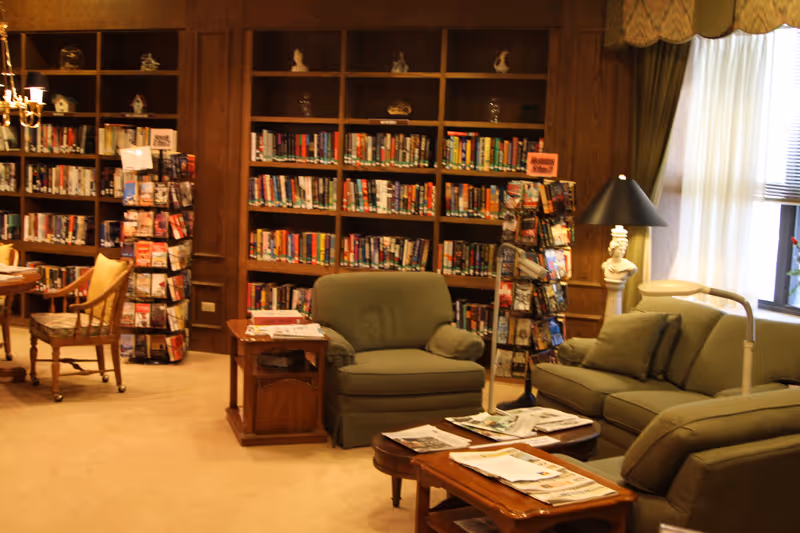 A cozy library or reading room with wooden bookshelves filled with books and decorative items. There are green upholstered armchairs and a sofa arranged around a wooden coffee table with newspapers on it. A wooden side table with more newspapers is next to one armchair. A floor lamp and a bust sculpture are near a window with curtains allowing natural light to enter.