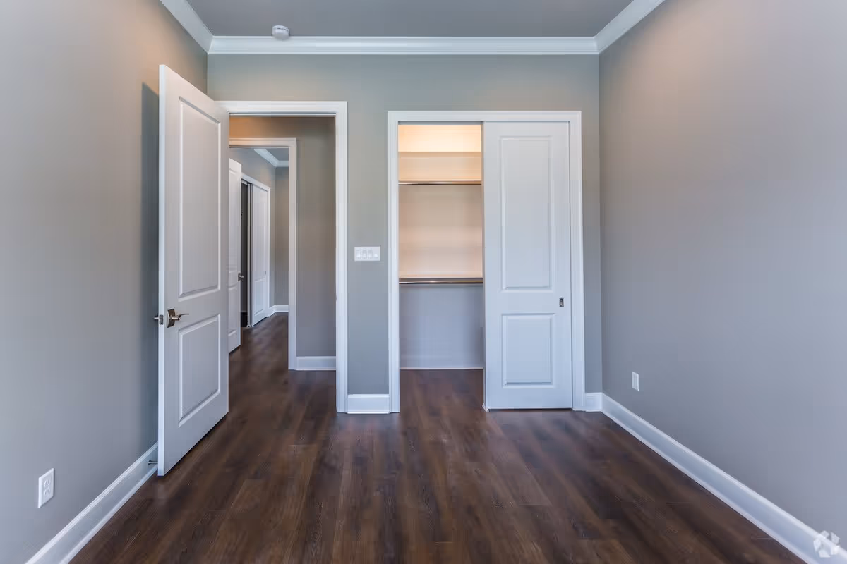 Empty room with gray walls, dark wood flooring, white baseboards, and crown molding. The room features an open white door leading to a hallway and a closet with sliding white doors, one of which is open showing a hanging rod inside.