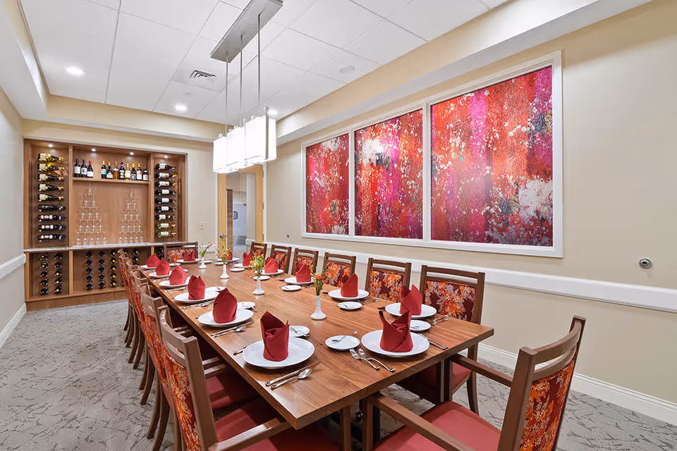 A formal dining room with a long wooden table set for twelve people. Each place setting has a white plate, silverware, a small side plate, and a red folded napkin. The chairs have red and orange floral upholstery. On the wall is a large three-panel abstract artwork in shades of red, pink, and white. At the far end of the room is a wooden wine rack filled with bottles and glasses. The room is well-lit with ceiling lights and a modern rectangular pendant light above the table.