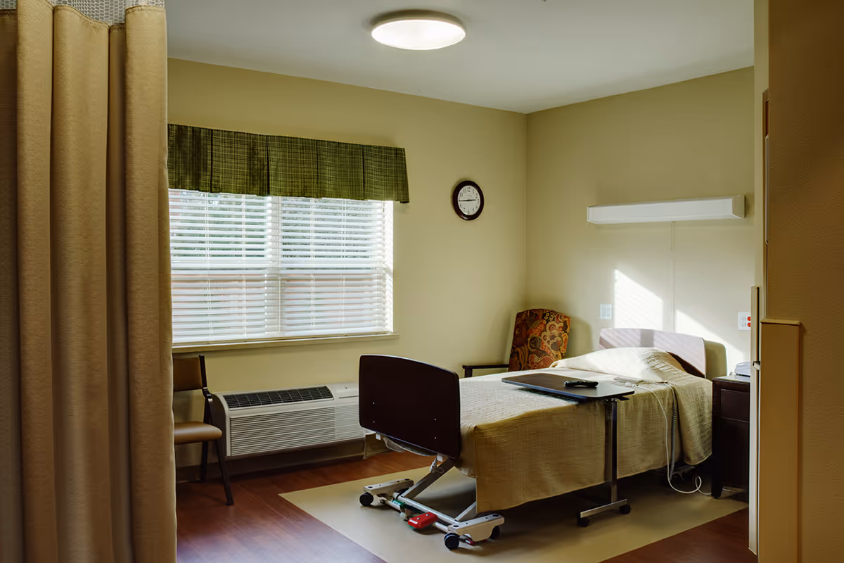 A senior living resident room with an adjustable bed, overbed table, armchair, window with blinds, and a privacy curtain.