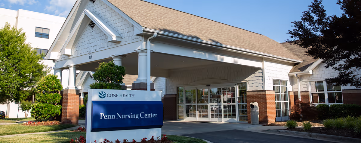 Exterior view of Cone Health Penn Nursing Center showing the entrance with a covered drop-off area, large windows, and a sign with the facility name in front. The building has white siding with brick accents and is surrounded by greenery and trees under a blue sky.