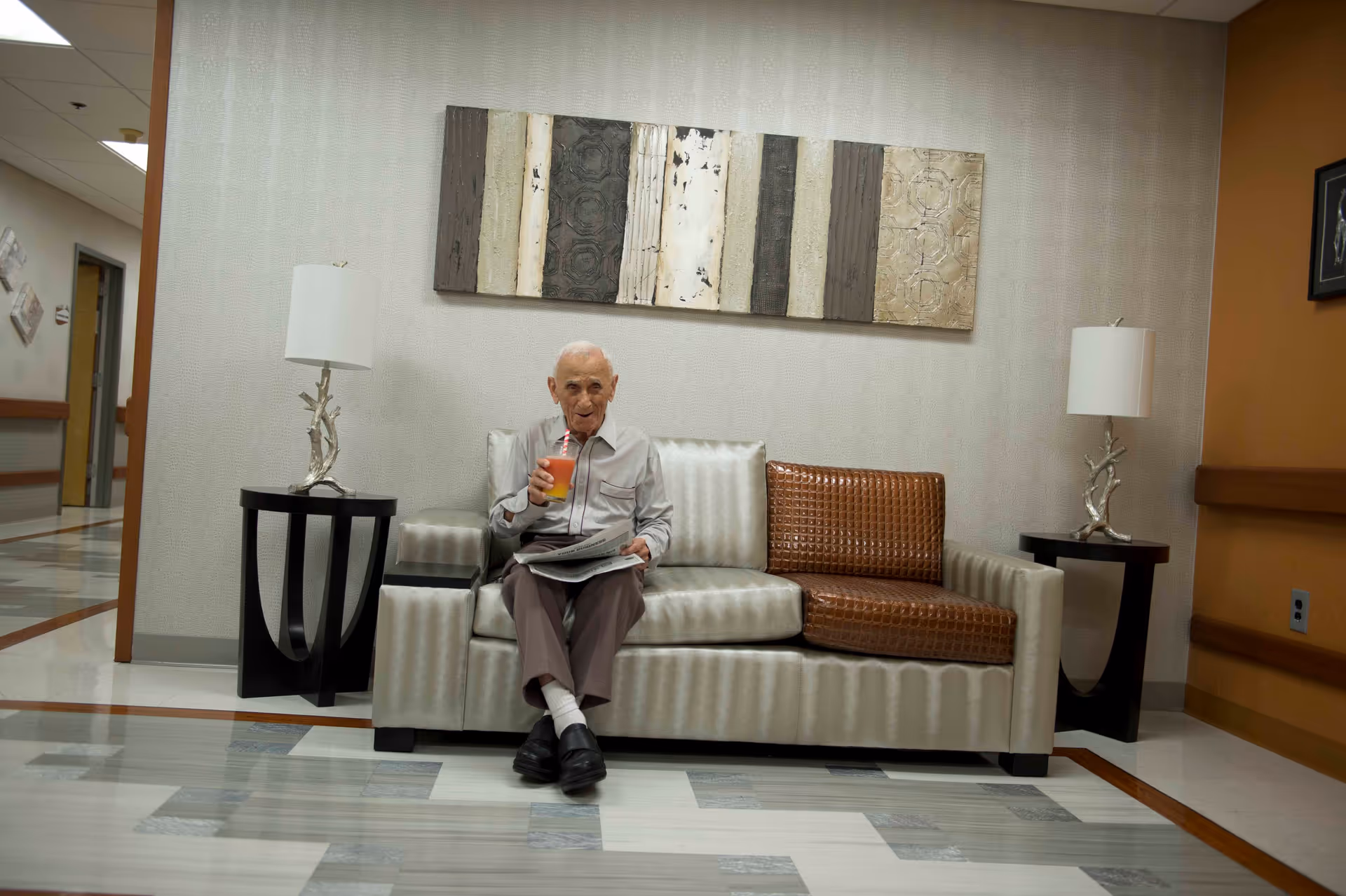 An elderly man sitting on a modern sofa in a hallway or common area of a healthcare facility, holding a drink and a newspaper. The sofa has a mix of silver and brown cushions. There are two side tables with lamps on either side of the sofa, and a decorative wall art piece hangs above it.