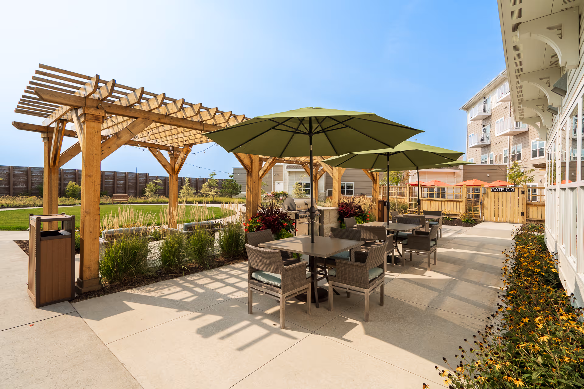 Outdoor patio area at Riley Crossing Senior Living featuring tables with green umbrellas, wicker chairs, a wooden pergola, and landscaped garden beds with flowers and grasses under a clear blue sky.