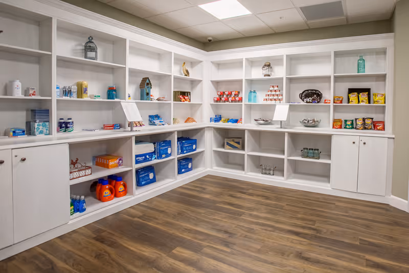 A corner of a room with white built-in shelves and cabinets stocked with various household and personal care items including laundry detergent, snacks, bottled drinks, and hygiene products. The room has wood flooring and a beige wall.