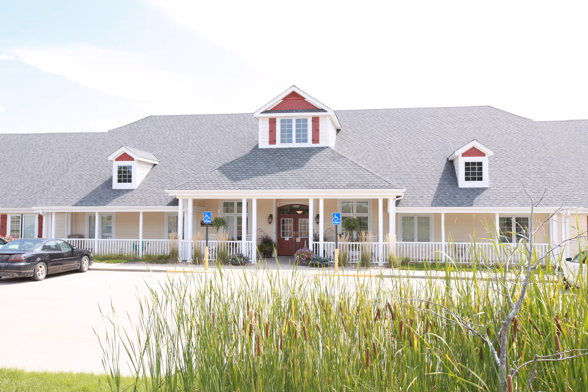 Front exterior view of a single-story assisted living facility building with a gray roof, white siding, and red accents around the windows and entrance. There is a covered porch with white railings, two handicap parking signs near the entrance, and some tall grass and plants in the foreground.