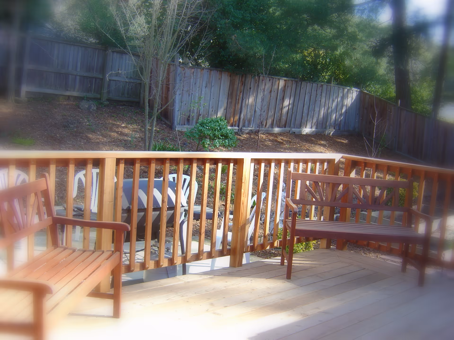 Wooden backyard deck with benches and a railing overlooking a fenced garden and trees.