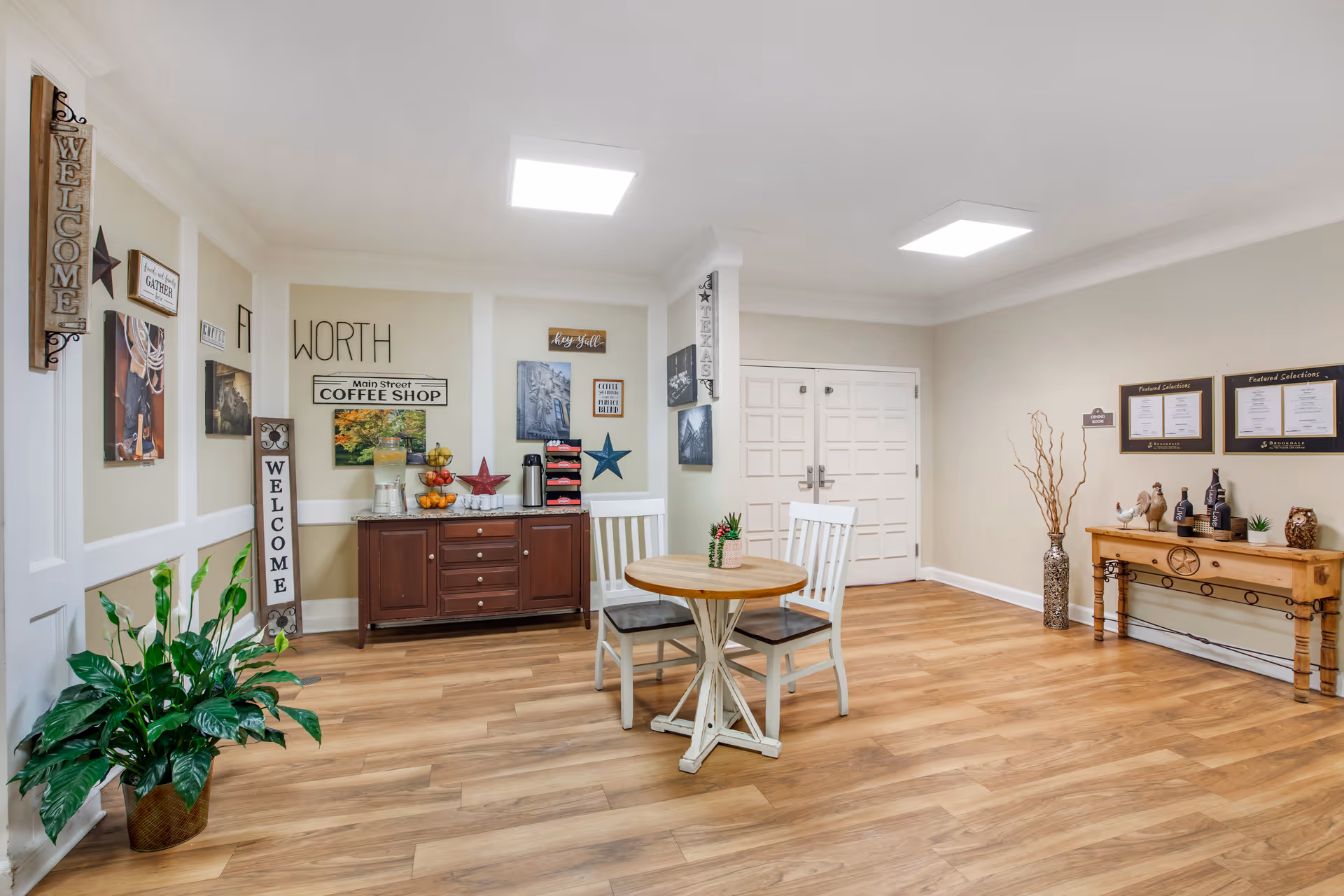 A bright and welcoming interior space with light wood flooring and beige walls. There is a small round wooden table with two white chairs in the center. Against the wall is a dark wooden cabinet with a water dispenser, fruit basket, coffee cups, and decorative stars. Various wall decorations include signs that say 'WELCOME', 'WORTH', 'Main Street COFFEE SHOP', and 'hey y'all'. On the right side, there is a wooden console table with decorative items and framed menus labeled 'Featured Selections'. A large green potted plant is on the left side near the entrance.