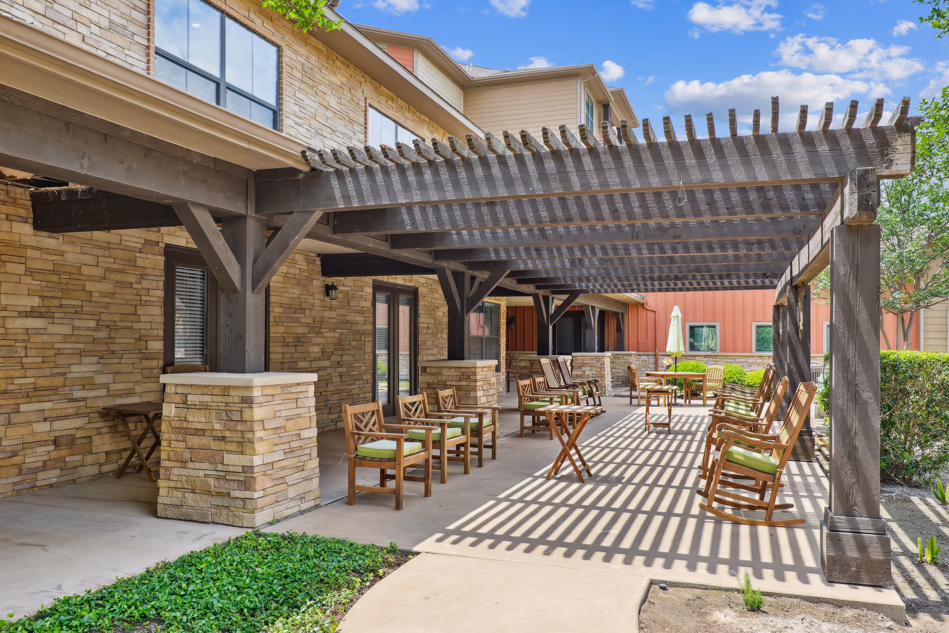 Outdoor patio area at Watercrest at Bryan with wooden pergola providing partial shade. Several wooden chairs with green cushions and small tables are arranged on the concrete floor. The building exterior features stone and siding walls, with windows and doors visible. Green shrubs and plants surround the patio area under a blue sky with some clouds.