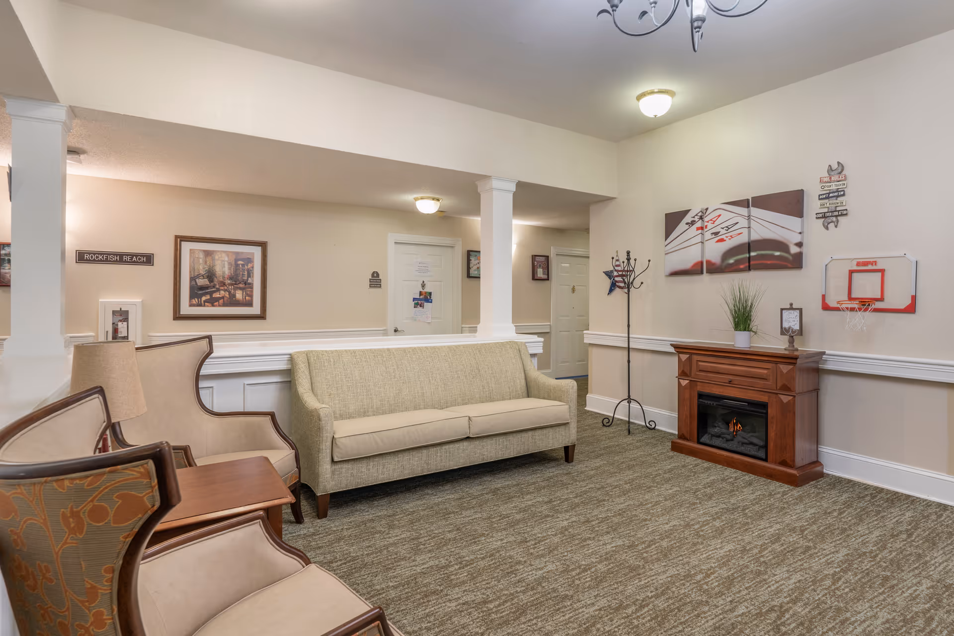 A cozy living room area in a senior living facility featuring a beige sofa, two armchairs with floral upholstery, a wooden side table, and a wooden electric fireplace with a small plant on top. The walls are light beige with white wainscoting, decorated with a three-panel artwork of playing cards and a small basketball hoop. There is a coat rack in the corner and several doors in the background.