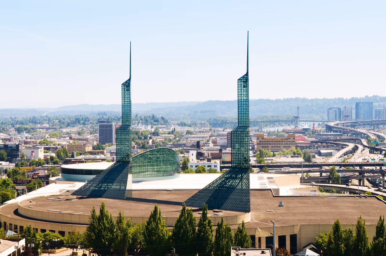 Aerial view of a large modern building with two tall glass spires casting shadows on the roof, surrounded by trees and urban infrastructure including highways and other buildings under a clear sky.