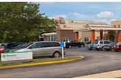 Exterior view of Ascension Living Villa Franciscan Place building with a parking lot in front, several parked cars, and a sign displaying the facility name near the entrance. Trees and a partly cloudy sky are visible in the background.