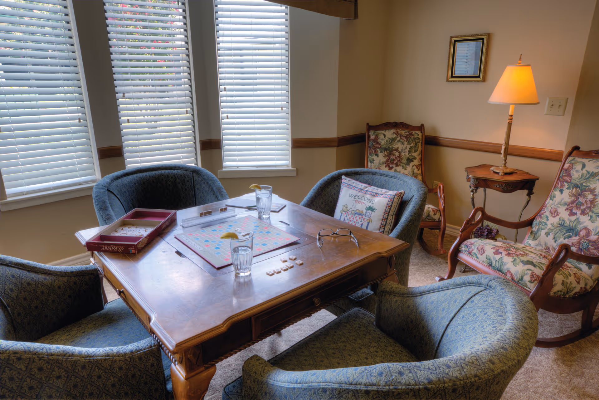 Cozy sitting area with a wooden table set up for Scrabble, surrounded by upholstered chairs and a lamp by windows with blinds.