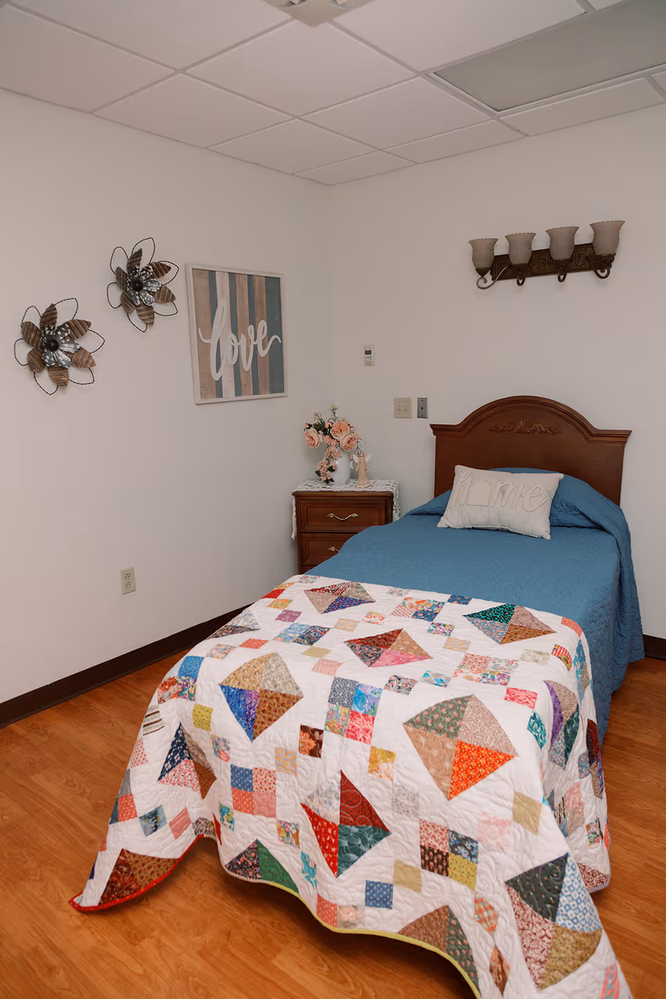 A cozy nursing home bedroom with a single bed covered in a blue bedspread and a colorful patchwork quilt. The bed has a pillow with the word 'Home' embroidered on it. Next to the bed is a wooden nightstand with a vase of flowers. The walls are decorated with two metal flower wall hangings and a framed artwork with the word 'love'. The room has a wooden floor and a ceiling with white tiles.