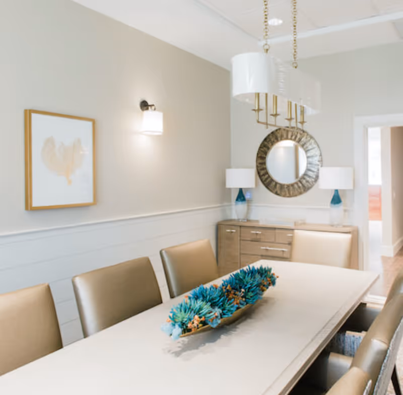Dining room with a long table and upholstered chairs, a blue-green centerpiece, sideboard with lamps and a round mirror under a pendant light.