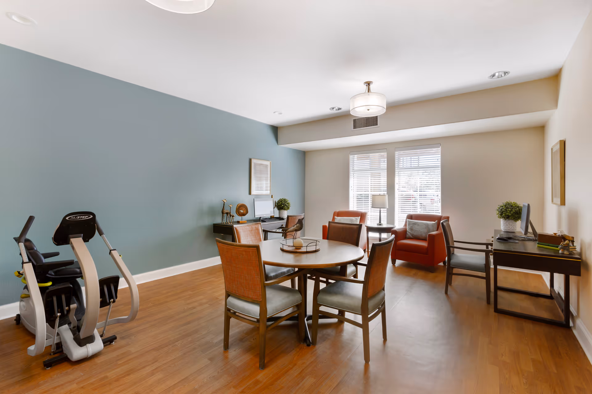 A bright room with wooden flooring featuring a round table surrounded by four chairs in the center. To the left, there is a NuStep exercise machine against a blue accent wall. At the back, two red armchairs flank a small round table with a lamp, positioned in front of two large windows with blinds. On the right side, there is a desk with a computer and a chair, and another chair nearby. The room has a modern ceiling light fixture and neutral-colored walls.