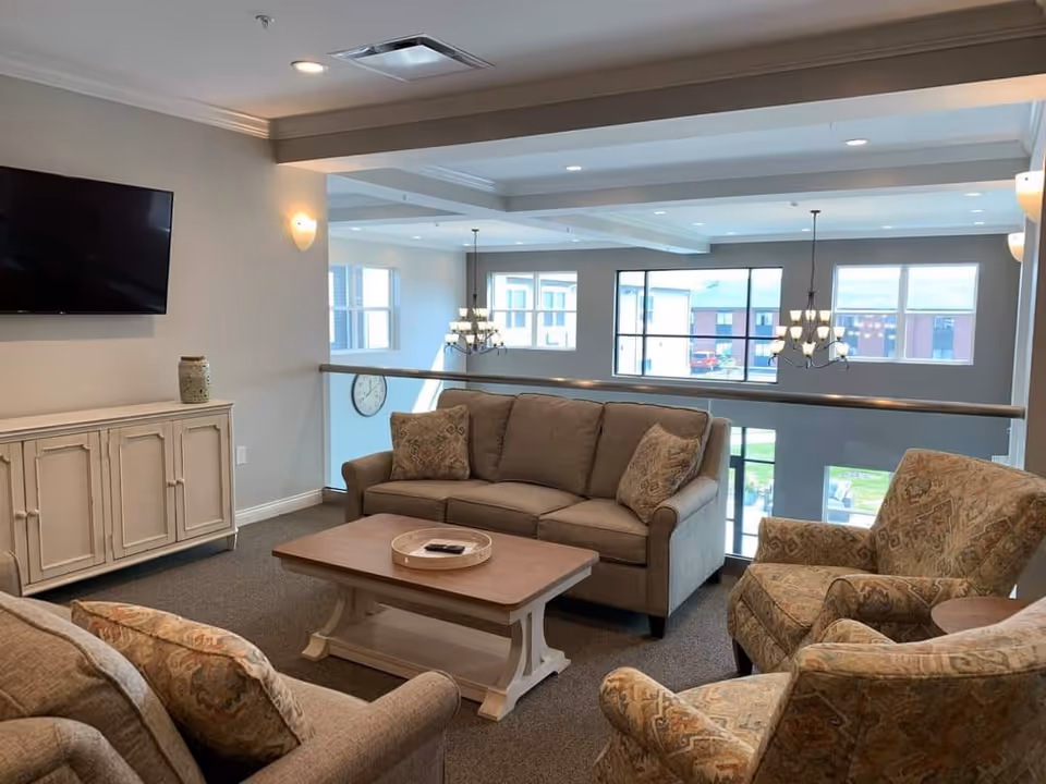 Bright communal living area with sofas and armchairs around a coffee table, a wall-mounted TV, and large windows overlooking the exterior.