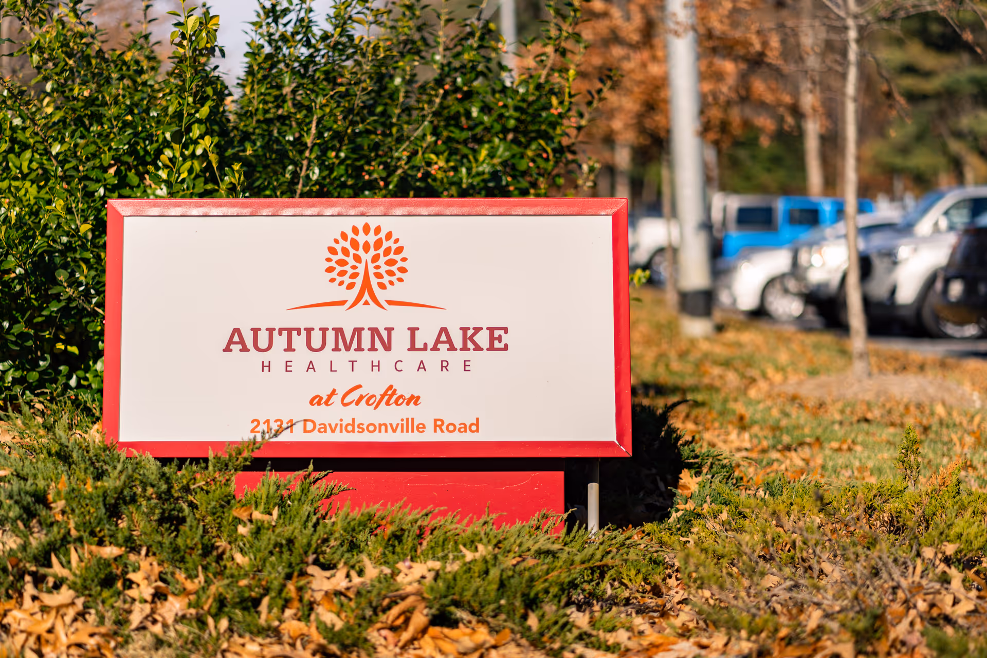A red and white sign for Autumn Lake Healthcare at Crofton located at 2131 Davidsonville Road, surrounded by greenery and fallen leaves with parked cars and trees in the background.
