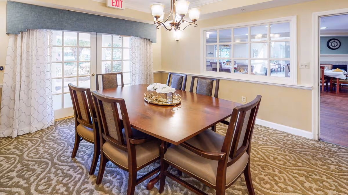 A dining room with a rectangular wooden table surrounded by eight cushioned chairs. The room has patterned carpet flooring, cream-colored walls, and a chandelier hanging above the table. There are large windows with white curtains and a glass door leading outside. A window on the right wall looks into another room with more tables and chairs.
