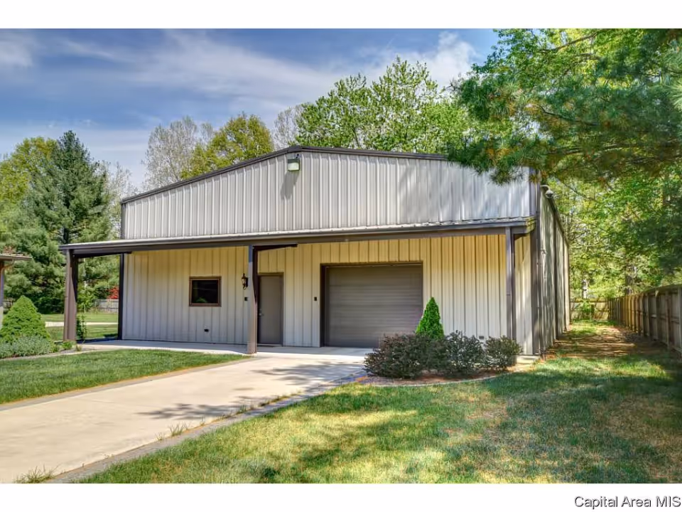 Exterior view of a metal building with a garage door and a small entrance door, surrounded by green grass, bushes, and trees under a partly cloudy sky.
