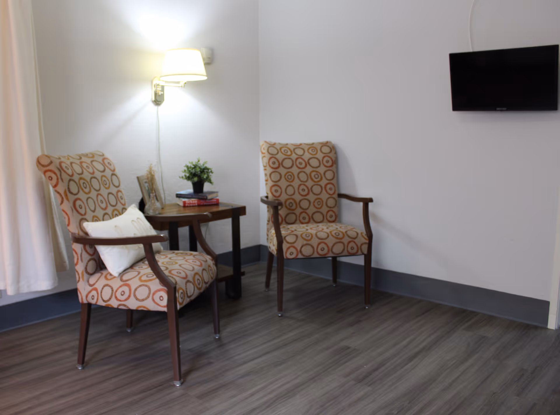 A small sitting area with two patterned armchairs and a wooden side table between them. The table holds a small potted plant, a picture frame, and some books. A wall-mounted lamp is above the table, and a flat-screen TV is mounted on the adjacent wall. The floor has wood-like vinyl flooring.